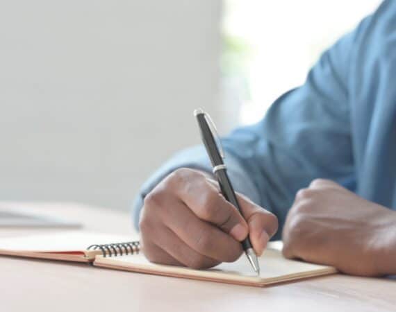 A man holding a pen, writing in a notebook while sitting at a desk.