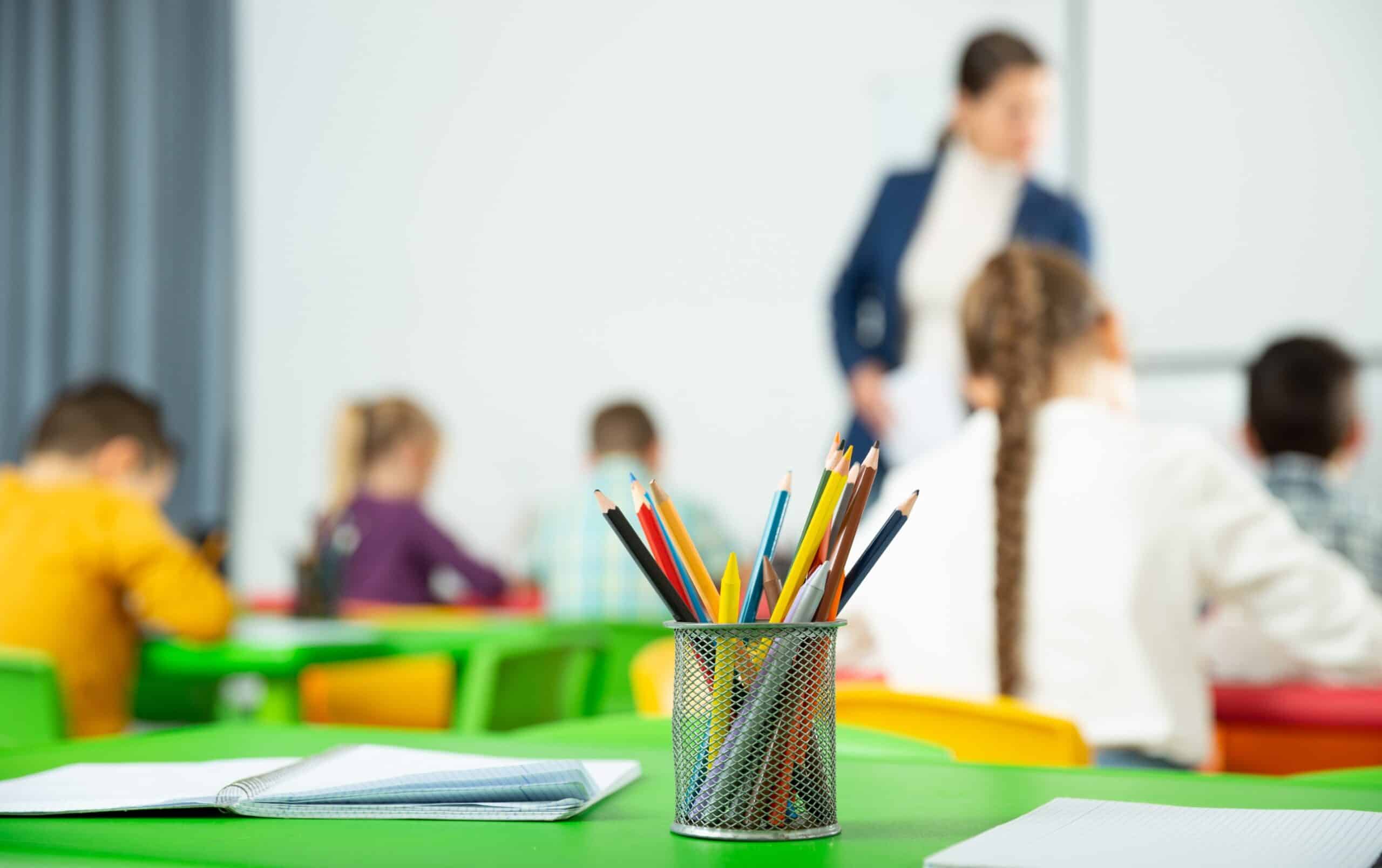 A cup of colored pencils on a table in a kindergarten classroom, while a designer is explaining UX to kids in the background.