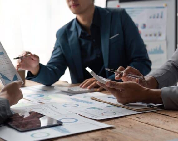 A group of business people sitting around a table with laptops and printed data reports.